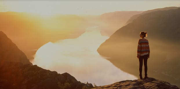 a woman standing near a cliff looking into a scenic valley. the sun is illuminating the body of water in the valley.