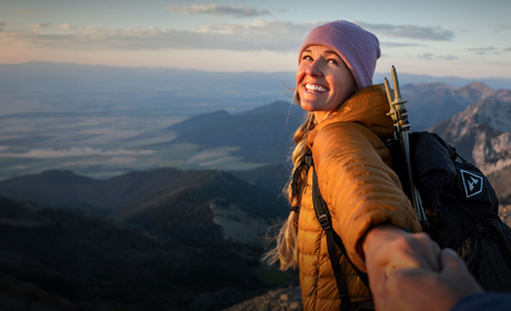 A woman smiling in a scenic park with mountains in the distance. She has her arm reaching out towards the camera