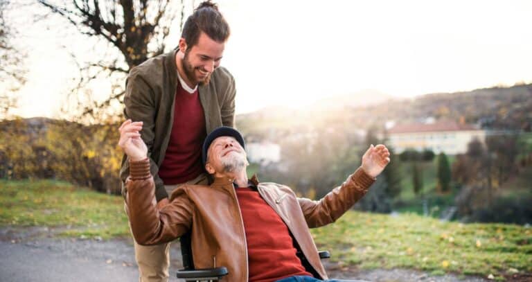 A younger man pushing an older man in a wheelchair. They are smiling and the sun is shining on them.